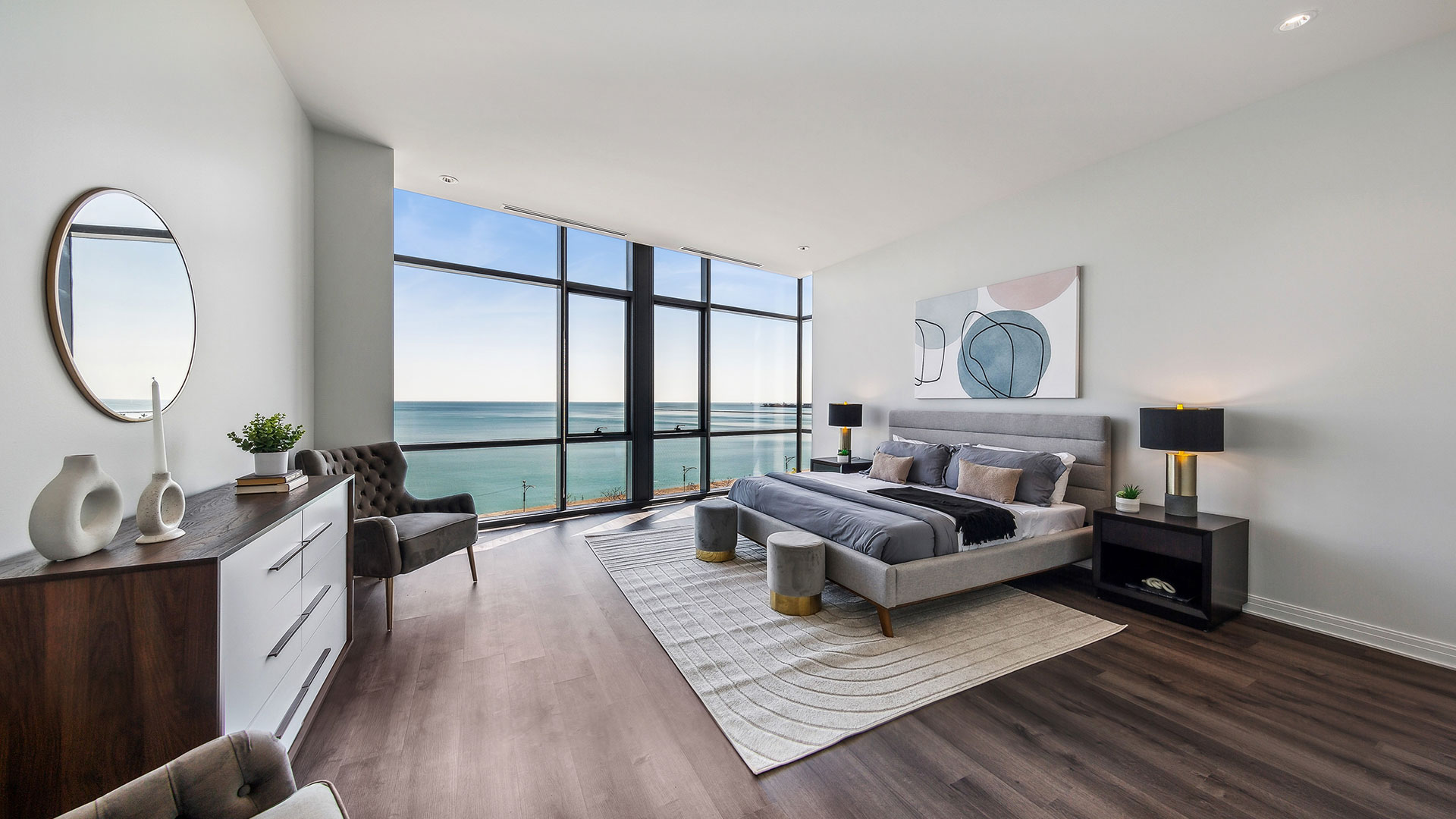 Modern bedroom with large floor-to-ceiling windows offering ocean views. Features a gray bed, black nightstands, white dresser, round mirror, and contemporary art on the wall.